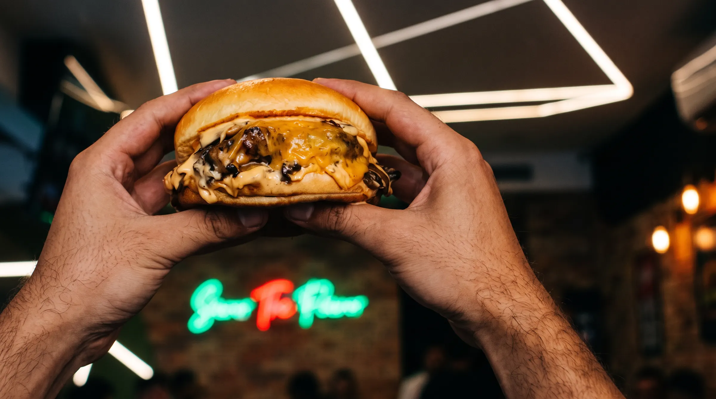 Hands holding a mushroom burger in front of the Savor The Flavor neon sign